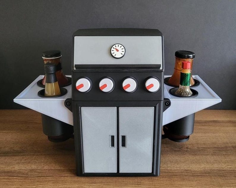 Puede incluir: Un soporte para condimentos en forma de mini-parrilla negra y gris con una parte superior y bandejas laterales plateadas. La parrilla tiene cuatro perillas rojas, un dial en forma de reloj y dos puertas plateadas. Dos botellas se encuentran en cada bandeja lateral.