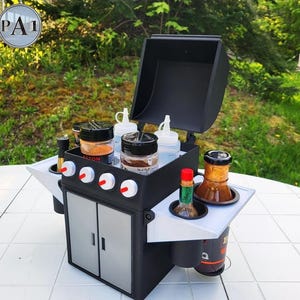 Puede incluir: Una peque&ntilde;a barbacoa negra y gris con detalles blancos, que contiene varios botes de especias, botellas exprimibles y botellas de salsa. La barbacoa tiene una tapa con bisagras y un armario con dos puertas. La imagen incluye el texto "PA1" en un c&iacute;rculo.