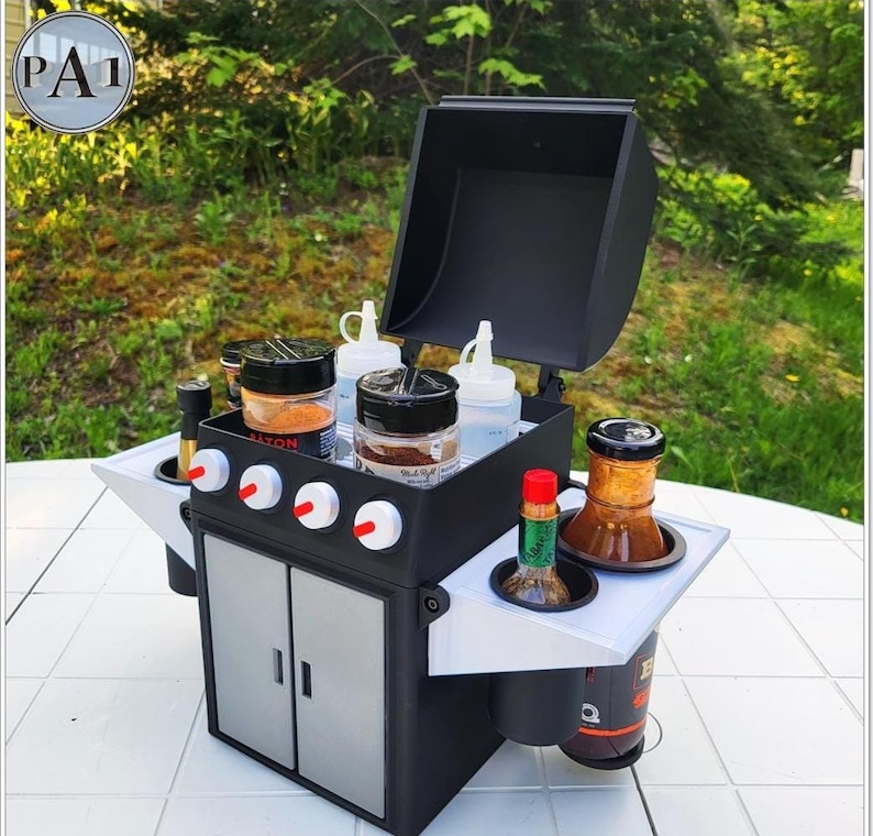 Puede incluir: Una peque&ntilde;a barbacoa negra y gris con detalles blancos, que contiene varios botes de especias, botellas exprimibles y botellas de salsa. La barbacoa tiene una tapa con bisagras y un armario con dos puertas. La imagen incluye el texto "PA1" en un c&iacute;rculo.