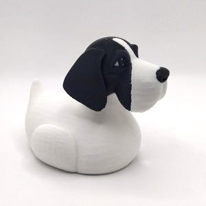 May include: A white and black dog figurine with a rounded body and a dog-like head. The dog has black ears and a black spot on its head. The nose is black, and the eyes are visible. The figurine is likely made of a solid material.