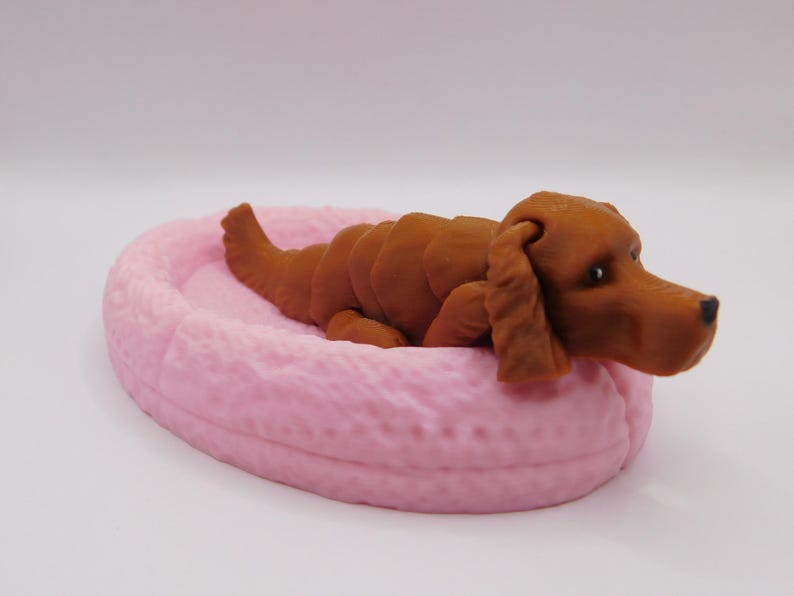 Op de afbeelding: Een bruin hondenbeeldje dat in een roze bed ligt. De hond ligt opgerold met zijn kop op zijn poten. Het bed is ovaalvormig en heeft een getextureerd oppervlak.