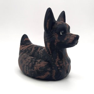 May include: A decorative figurine of a dog with a brindle pattern in shades of brown and black. The dog is in a seated position with its head turned to the right, ears perked up. The figurine is on a white background.
