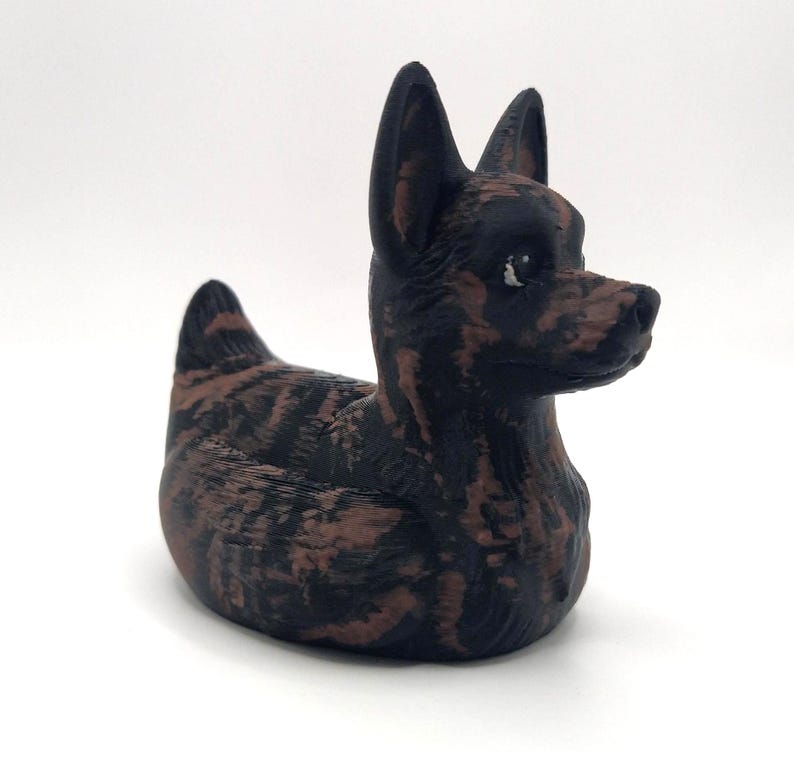 May include: A decorative figurine of a dog with a brindle pattern in shades of brown and black. The dog is in a seated position with its head turned to the right, ears perked up. The figurine is on a white background.