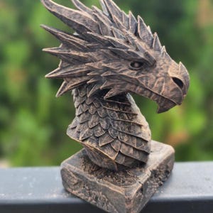 May include: A bronze-colored dragon bust sculpture. The dragon's head is detailed with textured scales, sharp spines, and a focused gaze. The sculpture is mounted on a square base, creating a striking decorative piece.