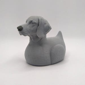May include: A gray dog figurine with a smooth finish. The dog has floppy ears, a pointed tail, and a black nose. The figurine is a decorative item, likely for home decor or a collector's item.