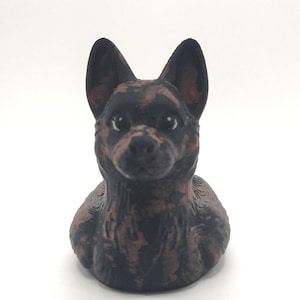 May include: A 3D-printed dog figurine in a sitting position. The dog has a black and brown mottled coat, with large, upright ears. The figurine is set against a plain white background.