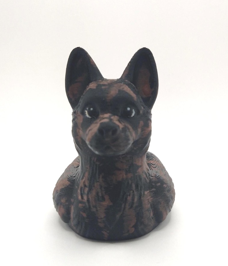 May include: A 3D-printed dog figurine in a sitting position. The dog has a black and brown mottled coat, with large, upright ears. The figurine is set against a plain white background.