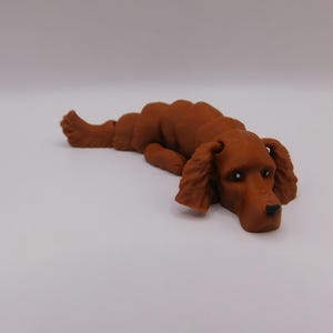 Op de afbeelding: Een bruin 3D-geprint beeldje van een hond die ligt met zijn hoofd op de grond. De hond heeft een lange snuit en slappe oren.
