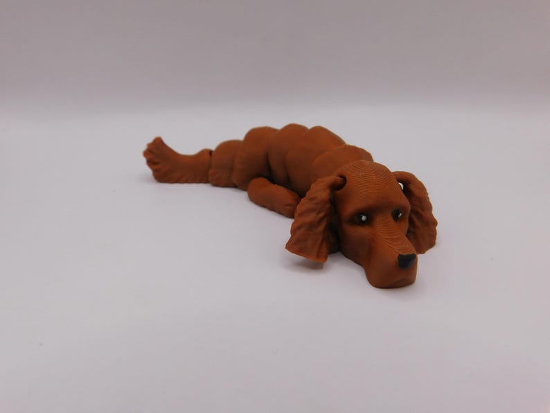 Op de afbeelding: Een bruin 3D-geprint beeldje van een hond die ligt met zijn hoofd op de grond. De hond heeft een lange snuit en slappe oren.