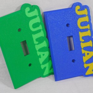 May include: Two light switch covers with the name "Julian" written in a retro font. One cover is green with a light green name and the other is blue with a yellow name.