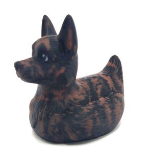 May include: A decorative figurine of a dog with a duck-like body. The dog is black and brown with a brindle pattern. The figurine has pointed ears and a small, rounded body. The dog is facing left.