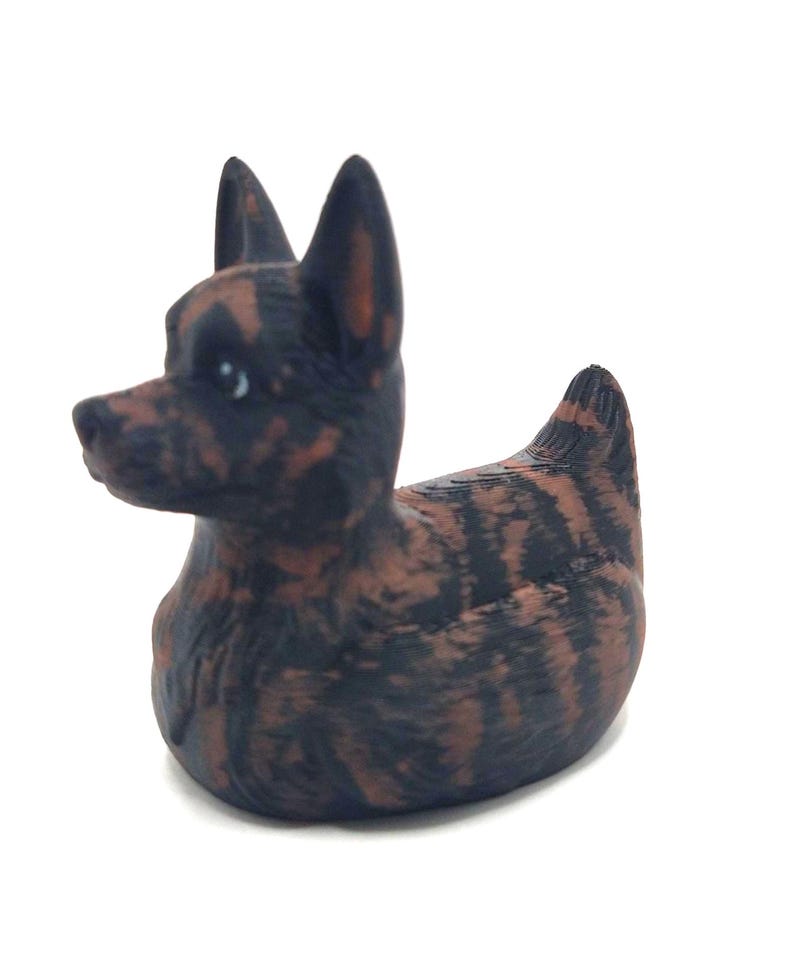 May include: A decorative figurine of a dog with a duck-like body. The dog is black and brown with a brindle pattern. The figurine has pointed ears and a small, rounded body. The dog is facing left.
