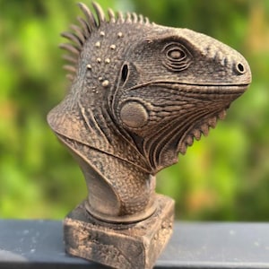 May include: A bronze-colored bust of an iguana, featuring detailed textures and scales. The sculpture has a textured base and a spiky crest along its head. The iguana's eye and facial features are intricately carved, showcasing a realistic reptile representation.