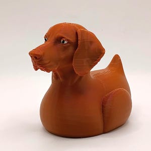 May include: A 3D-printed, orange-brown dog-shaped figurine. The dog has a duck-like body, with a pointed tail and a detailed head featuring eyes, a snout, and floppy ears. The figurine is designed with visible layer lines.