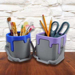 May include: Two gray paint can shaped desk organizers. One organizer holds paintbrushes and the other holds pencils and scissors. The organizers have a blue and purple rim.