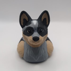 May include: A 3D printed figurine of a blue heeler dog. The dog is sitting with its head tilted slightly to the side. The figurine is painted in shades of gray, black, and brown.