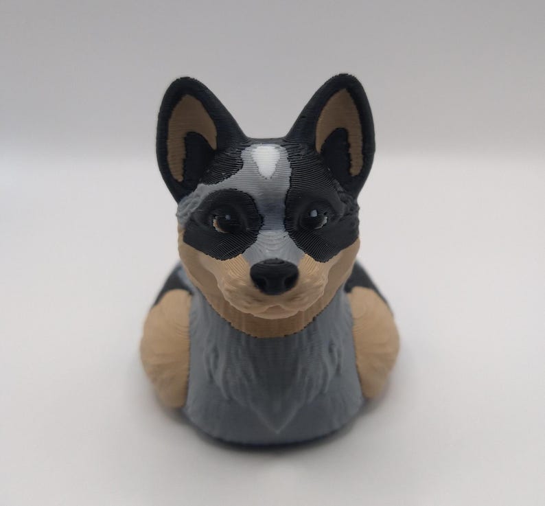 May include: A 3D printed figurine of a blue heeler dog. The dog is sitting with its head tilted slightly to the side. The figurine is painted in shades of gray, black, and brown.