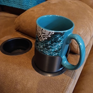 May include: A teal mug with a white and blue snowflake design sits in a black cup holder on a brown couch.