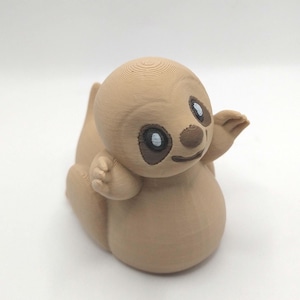 May include: A tan-colored, 3D-printed sloth figurine. The sloth has large, dark eyes with white highlights, a small brown nose, and a smiling mouth. Its arms are outstretched, and the body is rounded. The figurine is sitting down.
