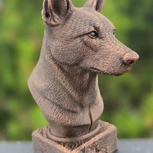 May include: A bronze-colored dog bust sculpture. The sculpture depicts the head and upper chest of a dog with pointed ears and a detailed textured surface. The dog bust is mounted on a square base.