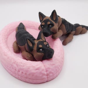 May include: Two black and brown German Shepherd dog figurines, one lying on a pink bed and the other lying on a white surface.