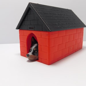 Op de afbeelding: Een rood en zwart plastic hondenhok met een baksteenpatroon. Een klein hondenhoofd kijkt uit de ingang.