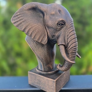 May include: Bronze-colored elephant bust sculpture. The sculpture features a detailed depiction of an elephant's head, including its large ears, trunk, and tusks. The bust is mounted on a rectangular base, creating a decorative piece.