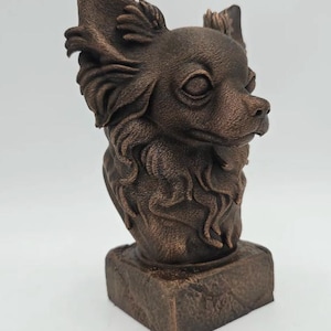 May include: A bronze-colored Chihuahua bust sculpture. The dog's head is detailed with fur texture, large ears, and a small nose. The sculpture sits on a square, textured base. The overall design is a decorative piece.