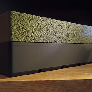 May include: A rectangular box consisting of two stacked sections. The top section features a textured, olive green surface, while the bottom section is a smooth, solid black base with a series of small, evenly spaced indentations along the bottom edge.