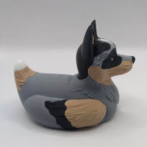 May include: A gray, black, and brown rubber duck shaped like an Australian Shepherd dog. The duck has a white patch on its chest and a black nose.