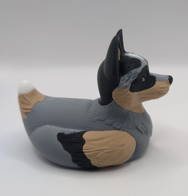 May include: A gray, black, and brown rubber duck shaped like an Australian Shepherd dog. The duck has a white patch on its chest and a black nose.