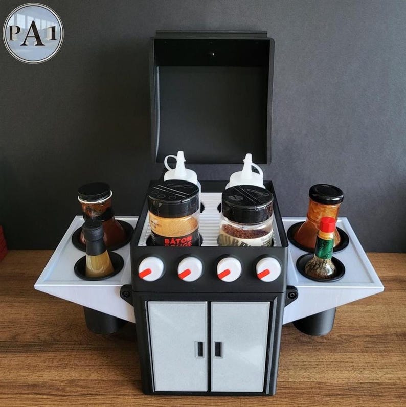Puede incluir: Un organizador de condimentos y especias con tem&aacute;tica de barbacoa en blanco y negro. El organizador tiene una tapa con bisagras, perillas y un dise&ntilde;o de puerta de armario. Contiene frascos de especias, botellas y botellas exprimibles. El texto "BATON ROUGE" es visible.