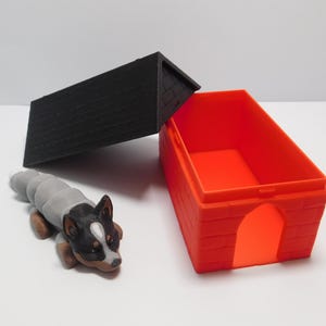 Op de afbeelding: Een klein, grijs en zwart hondenfiguurtje ligt naast een rood plastic hondenhok met een zwart plastic dak. Het dak is open en de binnenkant van het hondenhok is zichtbaar.