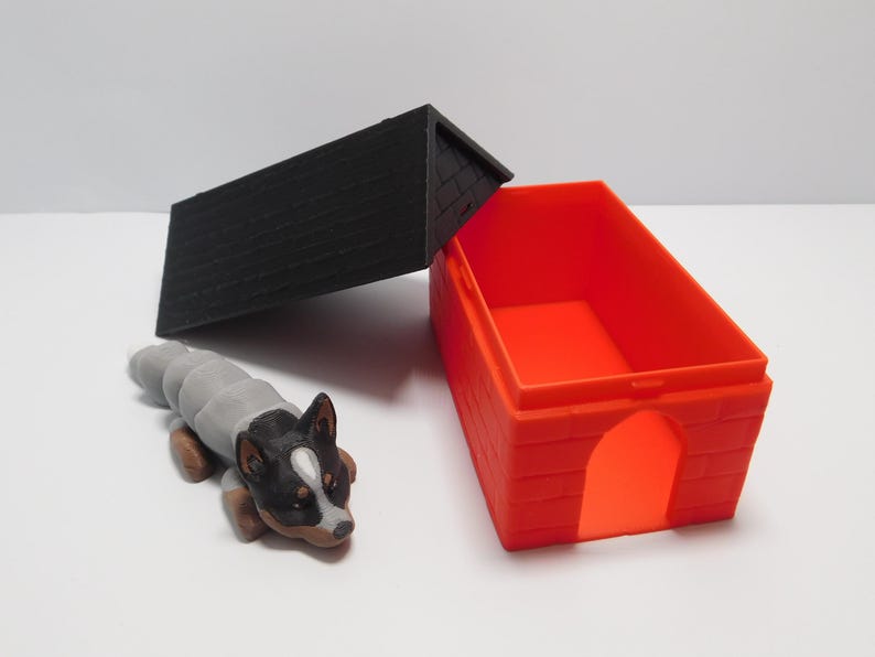 Op de afbeelding: Een klein, grijs en zwart hondenfiguurtje ligt naast een rood plastic hondenhok met een zwart plastic dak. Het dak is open en de binnenkant van het hondenhok is zichtbaar.