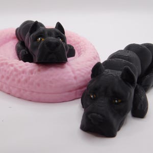 May include: Two black dog figurines, one lying in a pink dog bed and the other curled up. The dog figurines are detailed and realistic.