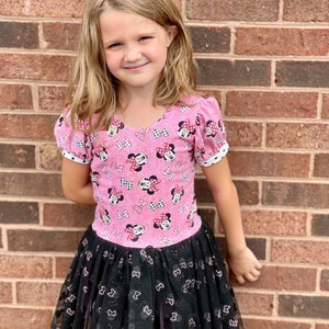 Puede incluir: Un vestido rosa y negro con estampado de Minnie Mouse. El vestido tiene una falda de tul y mangas cortas.