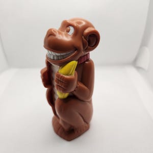 May include: A brown plastic monkey figurine holding a yellow banana. The monkey has a wide, toothy grin, white eyes, and a red collar around its neck. The figure is depicted in a standing position with a smooth, molded surface and rounded features.