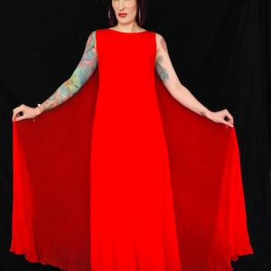 May include: A red maxi dress with a cape. The dress has a simple, sleeveless design and a scoop neckline. The cape is attached to the back of the dress and flows down to the floor.