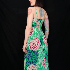May include: A green floral print dress featuring large pink and red roses, blue leaves, and white accents. The dress has thin straps and a fitted bodice. The dress is knee-length and made of a lightweight fabric.