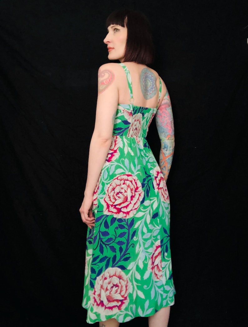 May include: A green floral print dress featuring large pink and red roses, blue leaves, and white accents. The dress has thin straps and a fitted bodice. The dress is knee-length and made of a lightweight fabric.