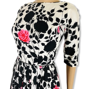 May include: A vintage dress featuring a floral pattern. The dress has a white background with black floral silhouettes and pink rose accents. It has a boat neckline, three-quarter sleeves, and a fitted bodice that flows into a pleated skirt.