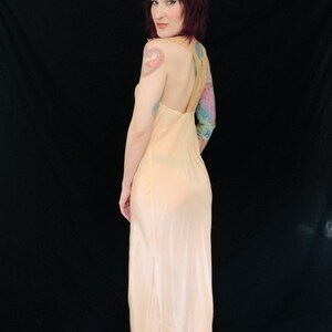 May include: A pale peach colored satin slip dress with a low back and spaghetti straps. The dress is floor length and has a slight sheen.