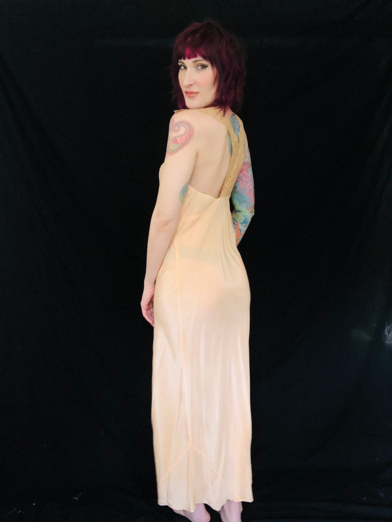 May include: A pale peach colored satin slip dress with a low back and spaghetti straps. The dress is floor length and has a slight sheen.
