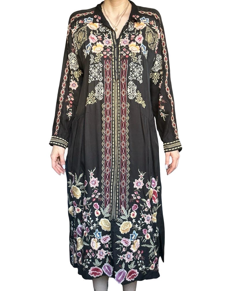 May include: A black long-sleeved midi dress featuring a central vertical panel with geometric patterns and colorful floral embroidery. The garment includes a collared neckline, button-down front, and intricate multi-colored flower motifs.