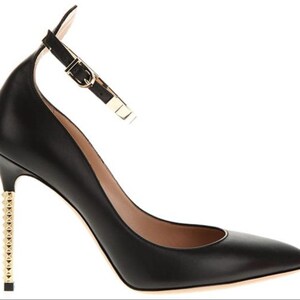 May include: Black leather high-heeled shoe with a pointed toe and ankle strap. The heel is embellished with gold-colored studs. The ankle strap features a gold-colored buckle. The shoe is designed for formal occasions.