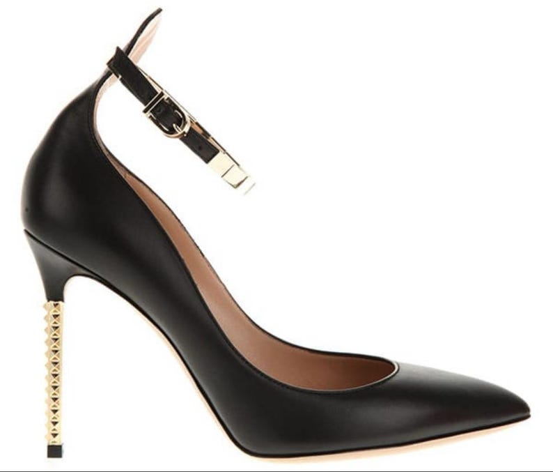 May include: Black leather high-heeled shoe with a pointed toe and ankle strap. The heel is embellished with gold-colored studs. The ankle strap features a gold-colored buckle. The shoe is designed for formal occasions.