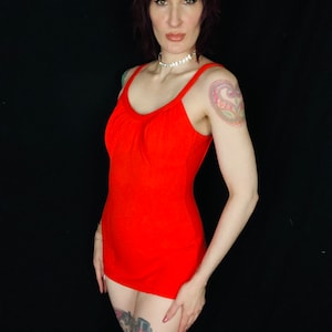 May include: A red one-piece swimsuit with a scoop neck and a fitted silhouette. The swimsuit has a simple design and is made of a smooth, stretchy fabric.