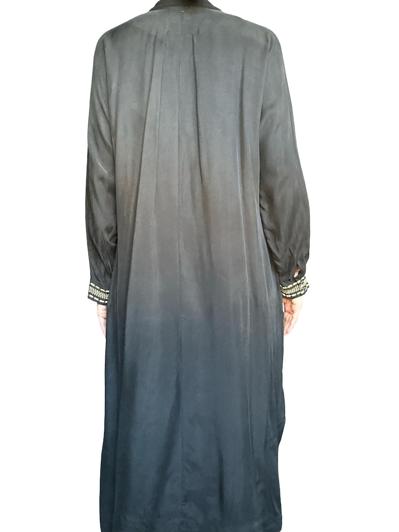 May include: A long-sleeved black duster coat featuring a subtle ombre effect that transitions to a darker shade at the hem. The garment includes long sleeves with decorative cuffs adorned with intricate gold and black beaded embroidery details.