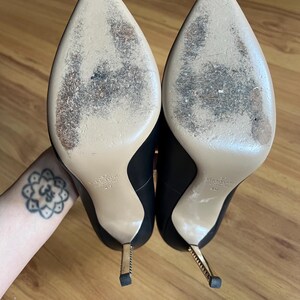 May include: A pair of black leather high-heeled shoes with pointed toes and gold-colored stiletto heels. The soles are beige with wear marks. The heels are approximately 4 inches tall.