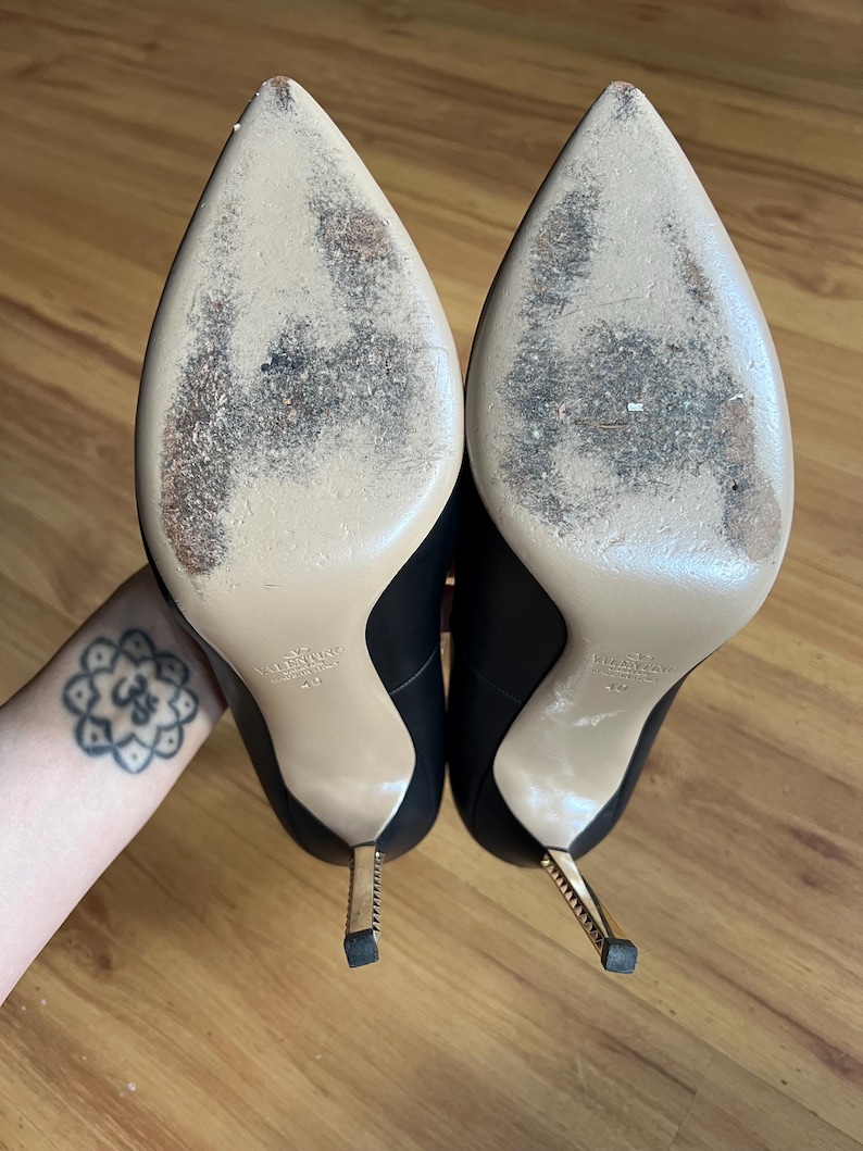May include: A pair of black leather high-heeled shoes with pointed toes and gold-colored stiletto heels. The soles are beige with wear marks. The heels are approximately 4 inches tall.